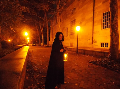 A hooded person carries a lantern on a lonely street.